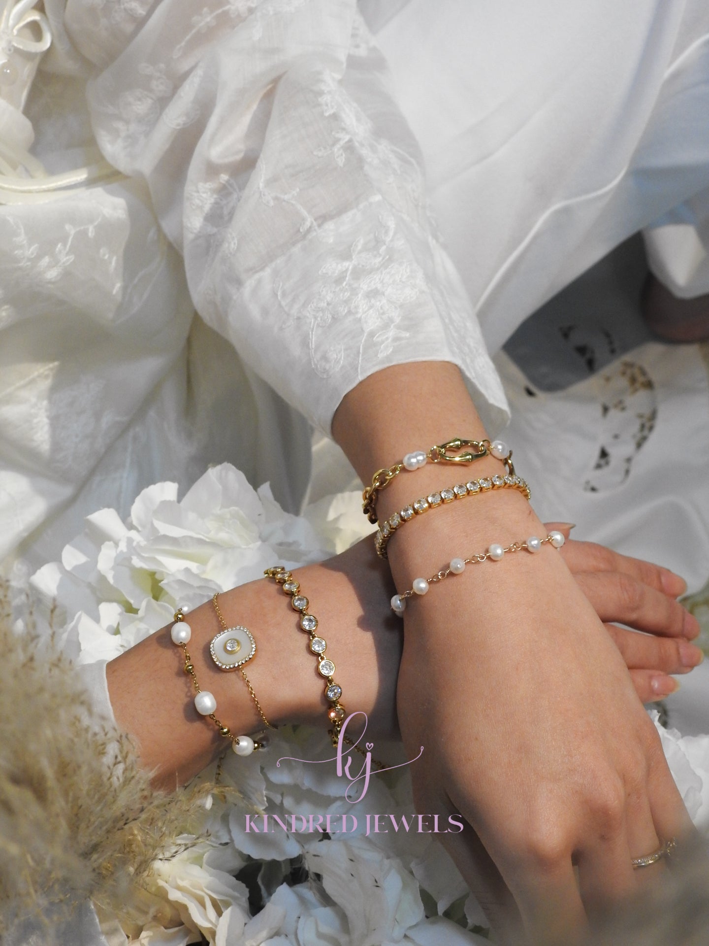 Close-up of hands wearing multiple pearl and gold bracelets on a white floral background with 'Kindred Jewels' branding.