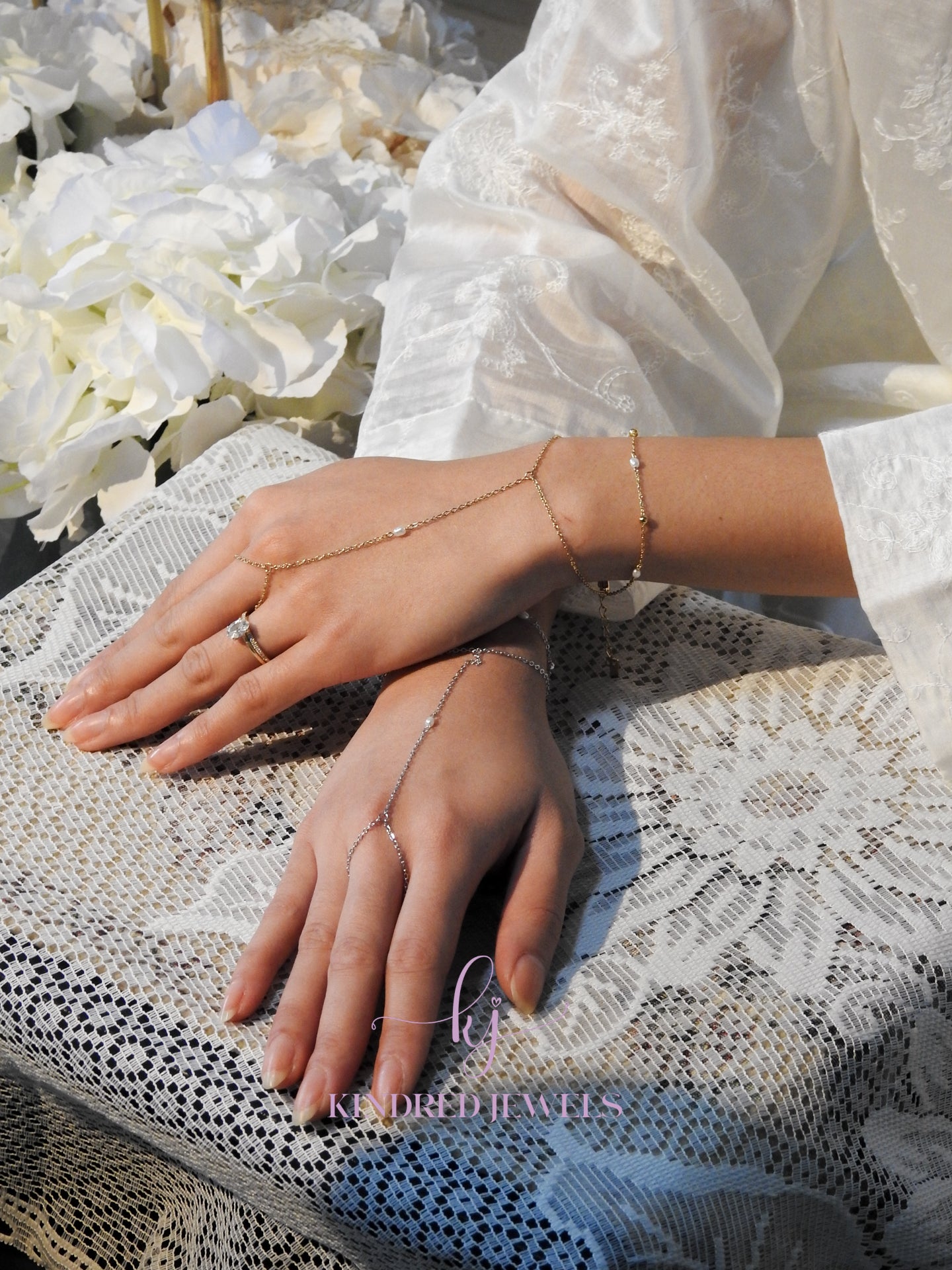 Close-up of hands wearing delicate jewelry on a textured white surface with flowers in the background.